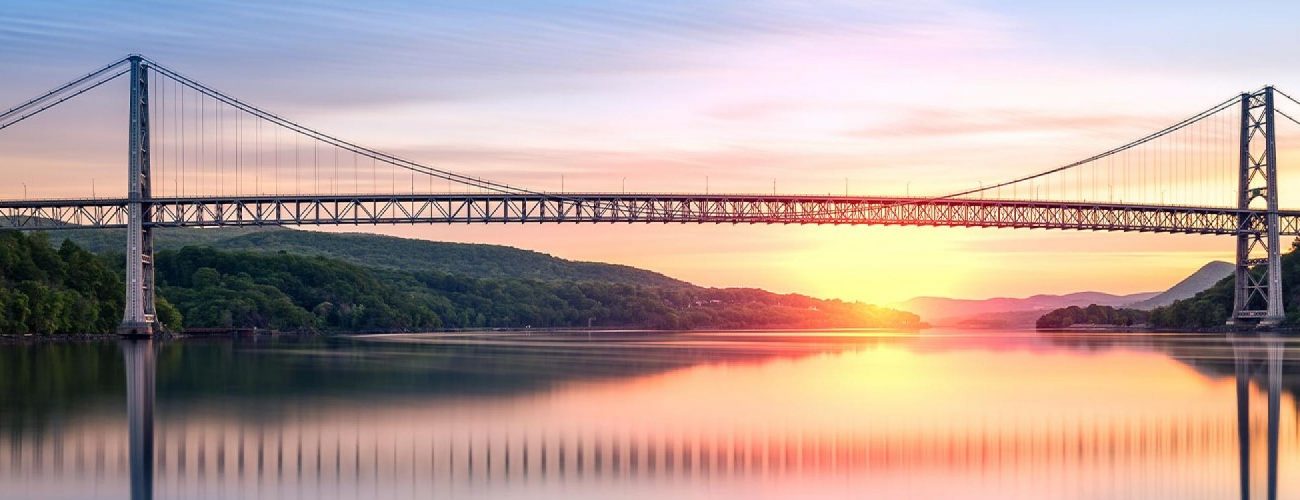 Suspension bridge spans a calm river at sunrise, with vibrant sky colors and lush green hills on both sides.