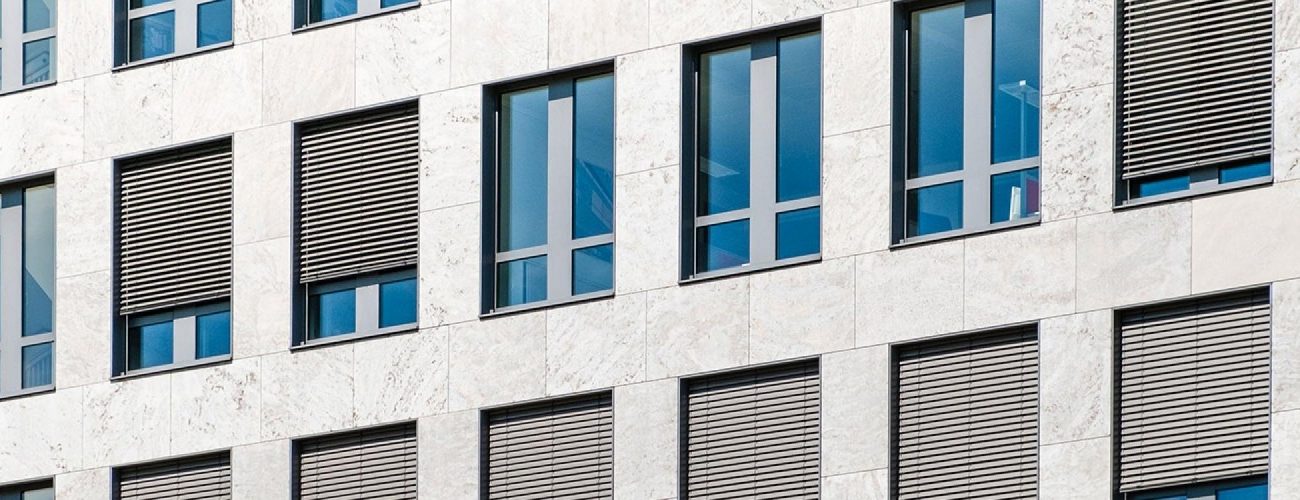 Modern building facade with alternating glass windows and closed gray shutters in a geometric pattern.
